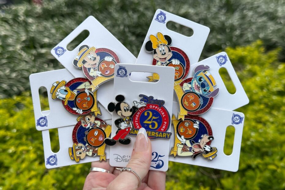 Hand holding a collection of six Disney 25th Anniversary pins featuring various characters, displayed in front of greenery.